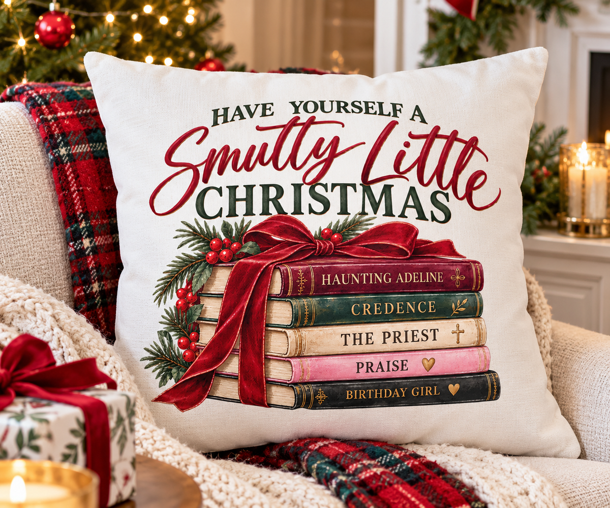 Have Yourself a Smutty Little Christmas book lover pillow styled on a green reading chair beside books and a holiday mug with a Christmas tree and fireplace behind it.