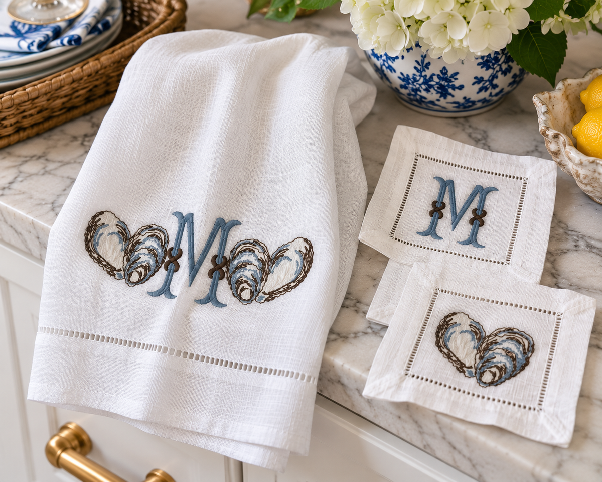 White 14 by 22 linen towel with oyster embroidery and blue monogram styled beside matching 6 by 6 cocktail napkins in a coastal kitchen.