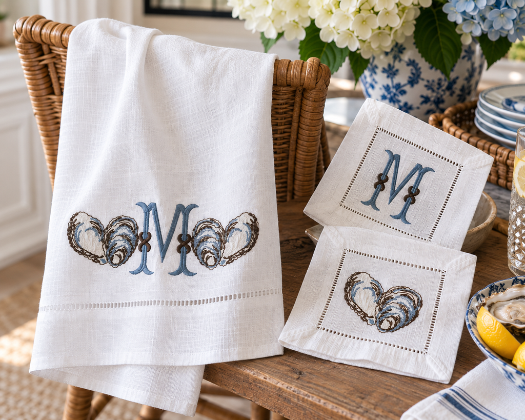 White linen towel with dusty blue monogram and embroidered oyster shells displayed with matching white hemstitched cocktail napkins featuring monogram and oyster designs.