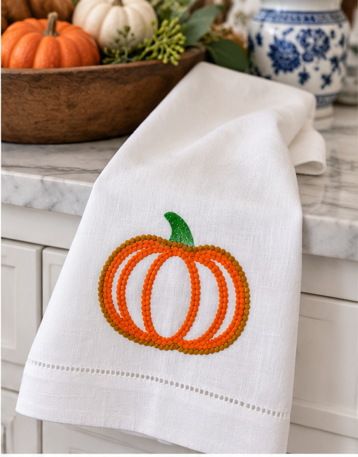 White linen fall kitchen towel displayed on a brass cabinet rail with dimensional orange embroidered pumpkin and green stem.