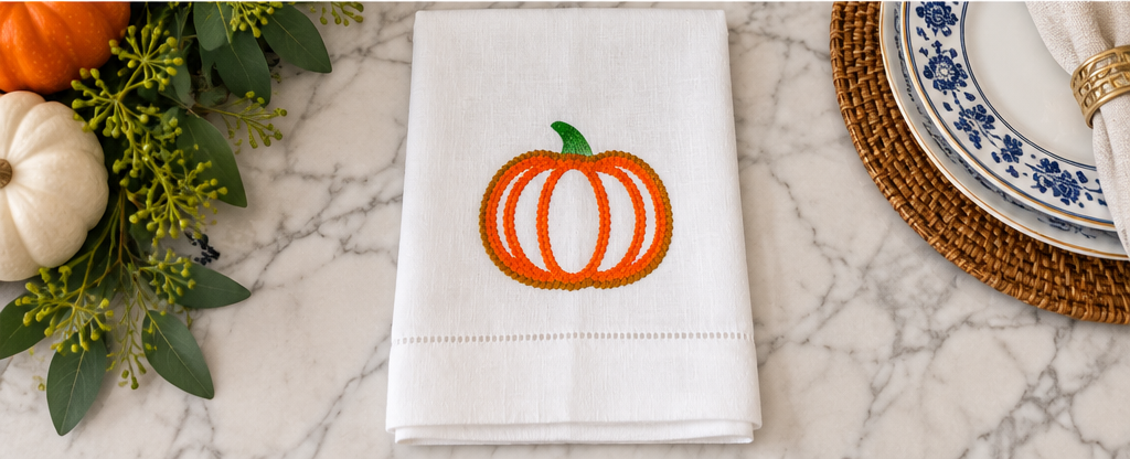 Textured white linen towel with orange and gold embroidered pumpkin displayed in a warm autumn kitchen setting.